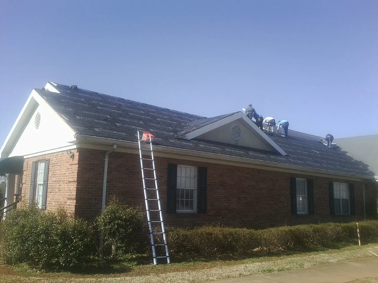 Crew tearing shingles off a brick ranch roof with a ladder set — Emergency Roof Repair work by our crew in Calverton