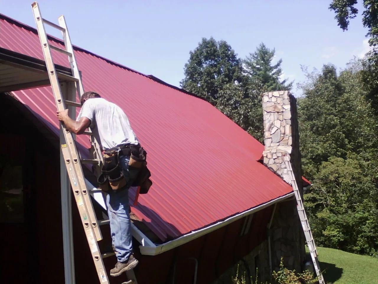 Roofer on a ladder beside a red metal roof and stone chimney — Soffit & Fascia Repair by Roofing Brothers in Calverton